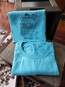 Our blue tee with a nopal that reads "chingona". The tee is folded and propped up on a box.