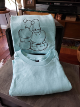 Load image into Gallery viewer, A closeup of our femme support illustrate on a light blue t-shirt. They are folded and leaning against a box.