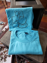 Load image into Gallery viewer, A closeup of our femme support illustrate on a blue t-shirt. They are folded and leaning against a box.