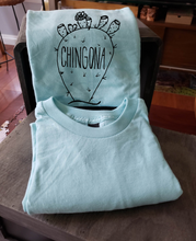 Load image into Gallery viewer, Our sky blue tee with a nopal that reads &quot;chingona&quot;. The tee is folded and propped up on a box.
