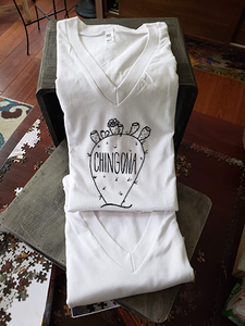 Our white v-neck tee with a nopal that reads "chingona". The tee is folded and propped up on a box.