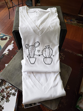 Load image into Gallery viewer, A closeup of our cacti holding hands on a v-neck white t-shirt. They are folded and leaning against a box.